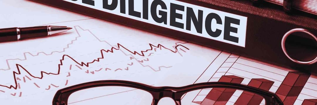 A due diligence binder rests atop financial charts displaying line and bar graphs, with a pen and a pair of glasses alongside.
