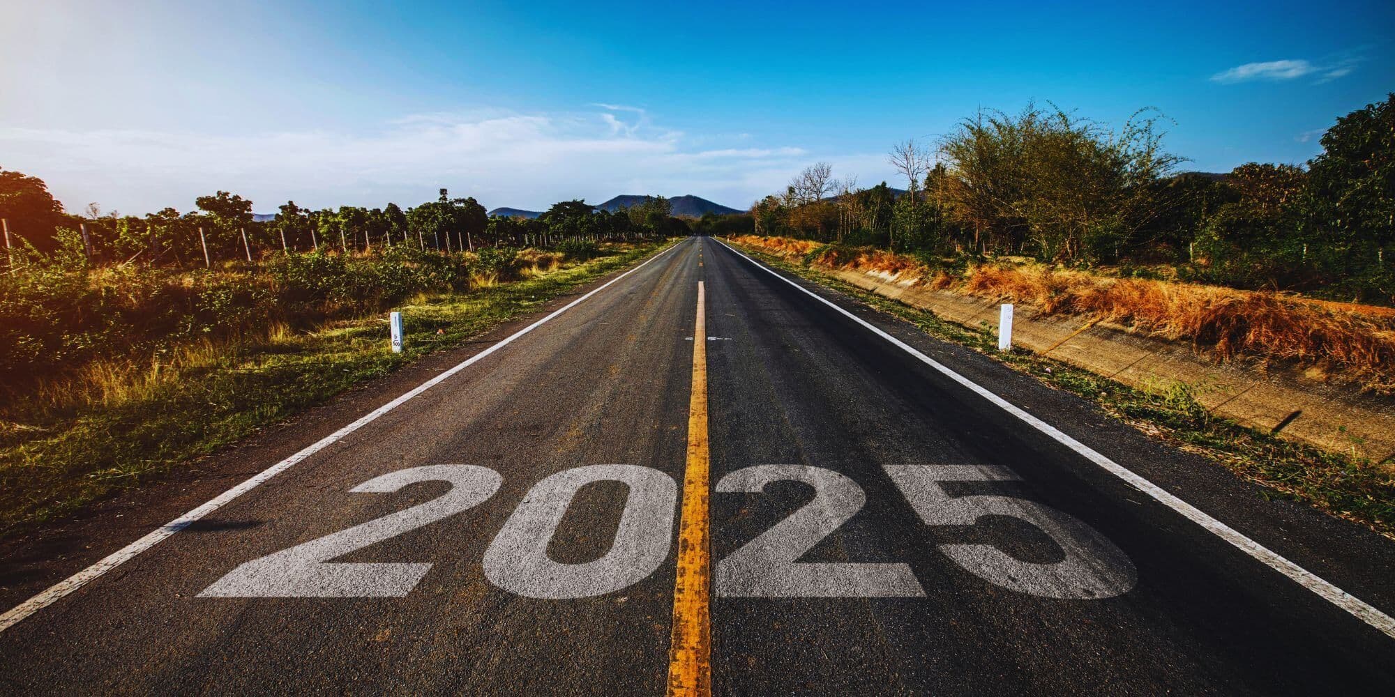 A straight country road stretching toward distant hills under a blue sky, with “2025” stenciled in large white numbers on the asphalt.
